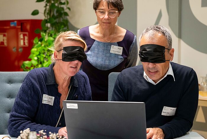 Eine Frau und ein Mann versuchen am Laptop, mit verbundenen Augen online ein Sofa zu bestellen.