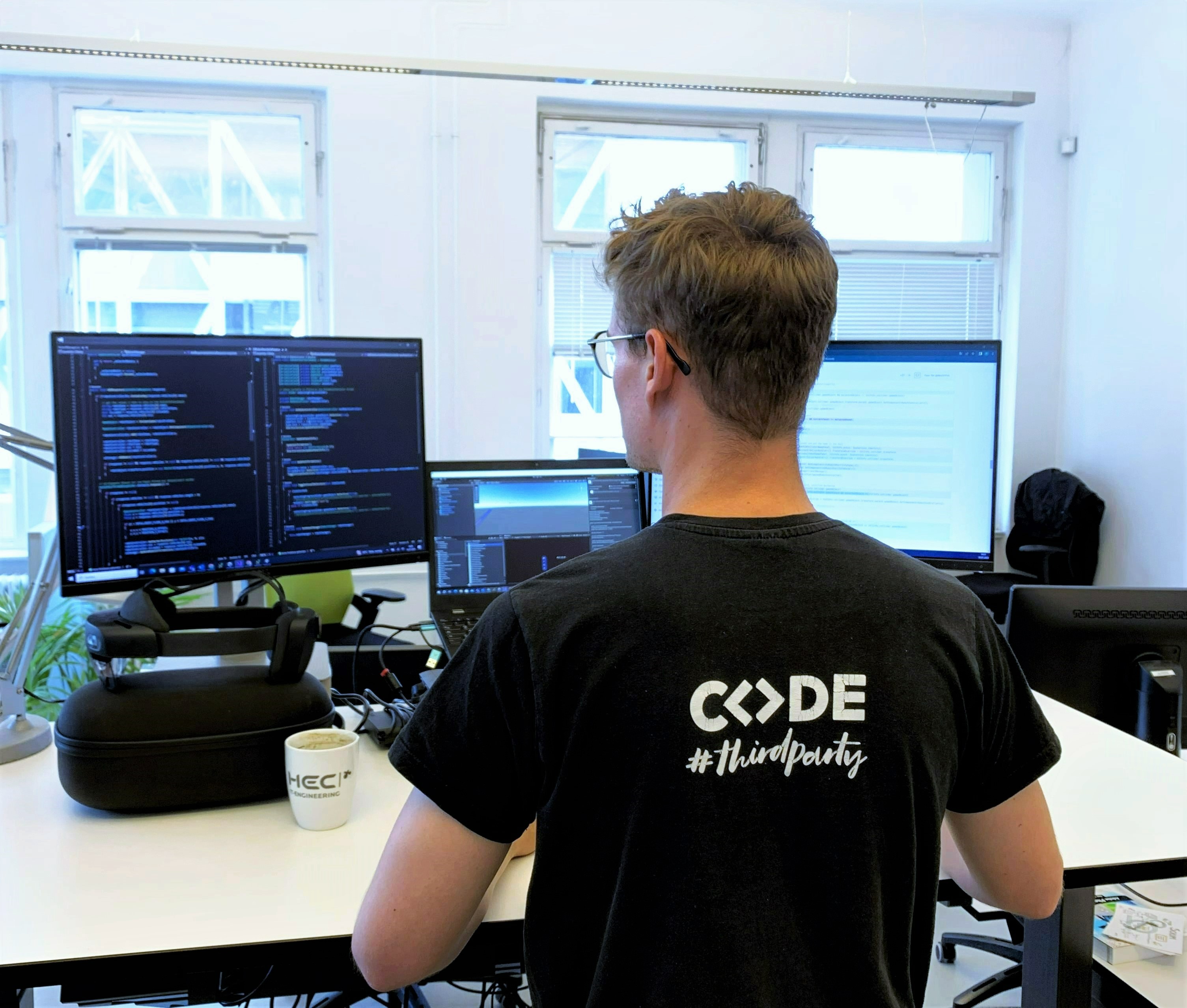 Paul Schilling, Werkstudent Werkstudent der HEC in Rückenansicht. Auf dem T-Shirt steht CODE. Er steht vor einem Schreibtisch.